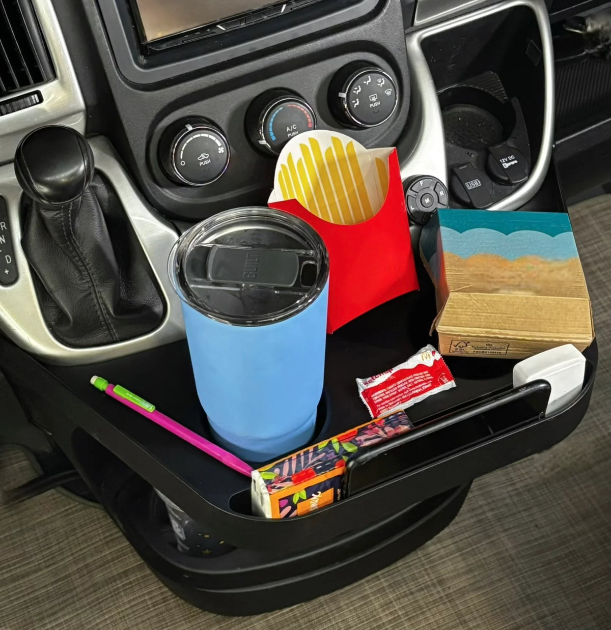 A promaster interior cup holder organizer tray holding a blue tumbler with lid, a red french fries bag, a small notebook, a pink pen, and other small items, shown installed around a gear shift console.