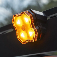 A close-up of a glowing amber diode dynamics scene light mounted on a black roof rack, with bright illuminated LED lenses and a blurred outdoor background.