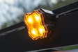 A close-up of a glowing amber diode dynamics scene light mounted on a black roof rack, with bright illuminated LED lenses and a blurred outdoor background.