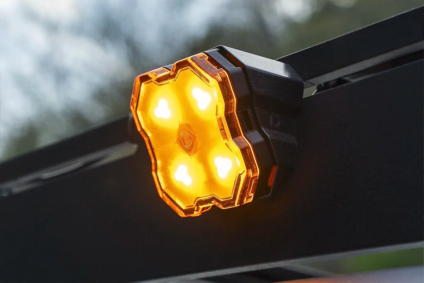 A close-up of a glowing amber diode dynamics scene light mounted on a black roof rack, with bright illuminated LED lenses and a blurred outdoor background.