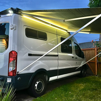 A white Ford Transit van parked outdoors in a backyard with Orion Van Gear roof rack and an extended Fiamma F45s awning and warm white LED strip lights attached above the side windows. The scene is set at dusk with grass, a wooden fence, and trees visible.