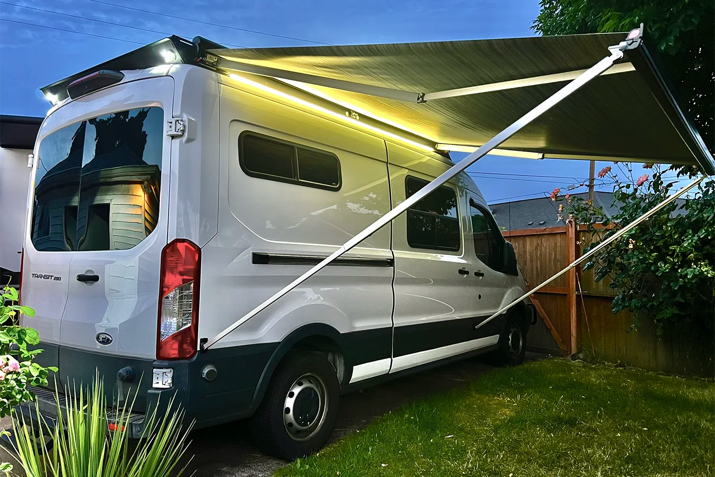 A white Ford Transit van parked outdoors in a backyard with Orion Van Gear roof rack and an extended Fiamma F45s awning and warm white LED strip lights attached above the side windows. The scene is set at dusk with grass, a wooden fence, and trees visible.