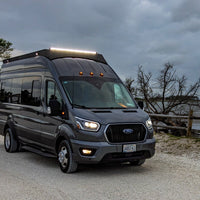 A dark gray Ford Transit recreational van parked on a gravel path outdoors near a body of water under an overcast sky and 50 inch lightbar on