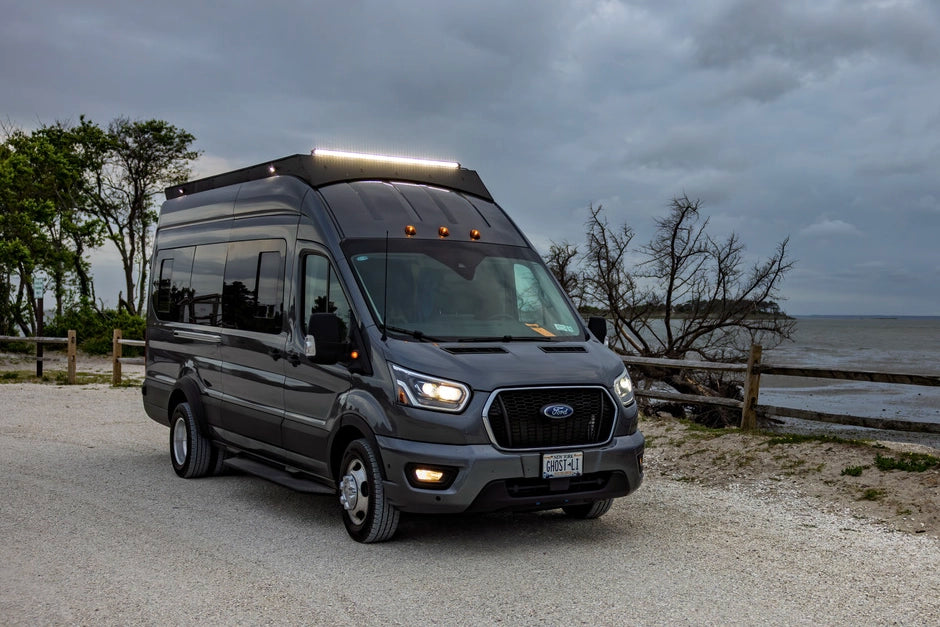 A dark gray Ford Transit recreational van parked on a gravel path outdoors near a body of water under an overcast sky and 50 inch lightbar on
