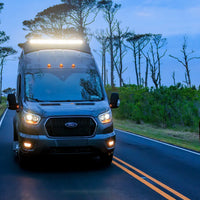A Ford transit van with bright headlights and roof lightbar driving on a rural road at dusk, with trees and horses visible on both sides of the road.