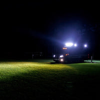 A dark vehicle with bright white and blue lights parked on a grassy field at night, casting a strong beam of light onto the lawn.