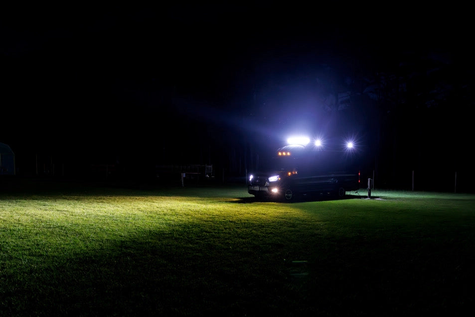 A dark vehicle with bright white and blue lights parked on a grassy field at night, casting a strong beam of light onto the lawn.