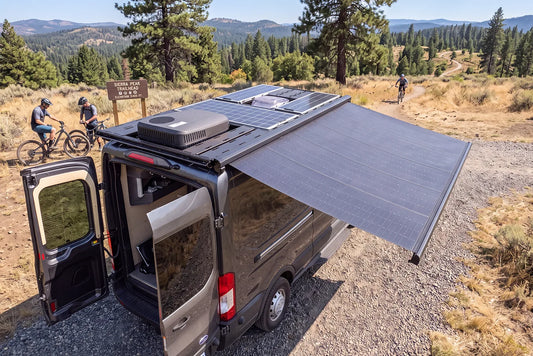 Orion Van Gear Ford Transit roof rack with extended solar awning, rooftop solar panels, and air conditioner at a scenic mountain biking trailhead.