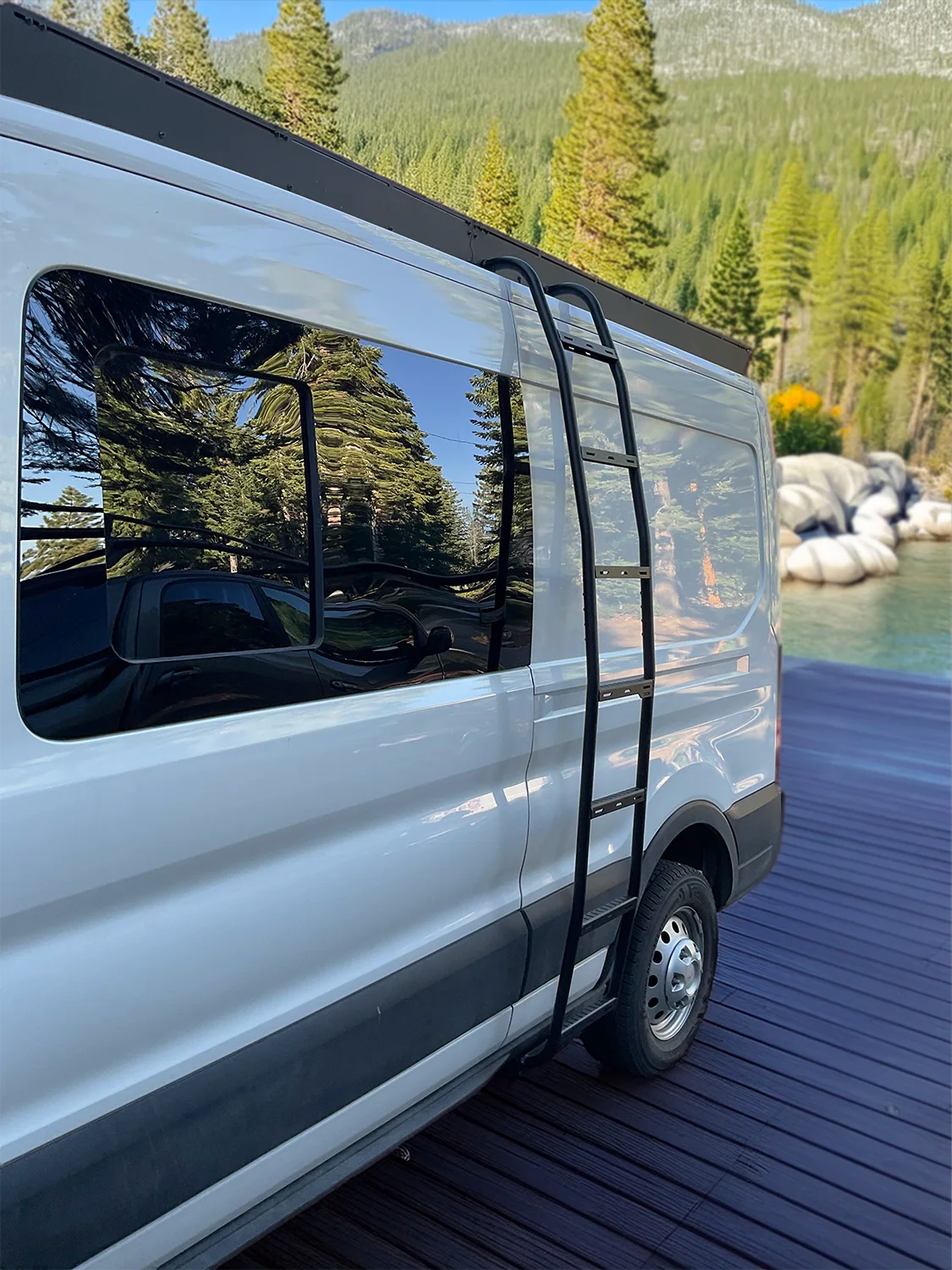 A white van parked on a dark wooden dock with a forest and mountains in the background. A black Orion Van Gear ladder is attached to the side of the van, and the windows reflect the surrounding trees.