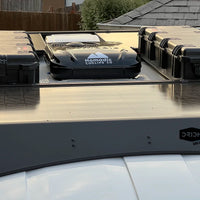 An Orion Van Gear Transit roof rack system carrying two black hard-sided storage cases on a vehicle roof, with flush-mounted solar panels and Nomadic X2 Air Conditioner