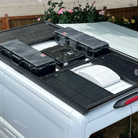 Black orion roof rack-mounted rich solar panels, air conditioner, and hard cases installed on the roof of a white mid-roof transit van, with a garden fence and plants visible in the background.
