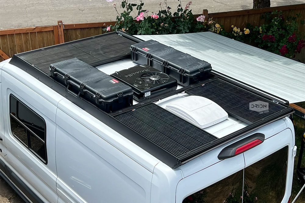 Black orion roof rack-mounted rich solar panels, air conditioner, and hard cases installed on the roof of a white mid-roof transit van, with a garden fence and plants visible in the background.
