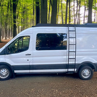 A white mid roof Ford Transit cargo van with orion van gear roof rack and side ladder parked on a dirt path in a forest with green trees and brown leaf litter on the ground.