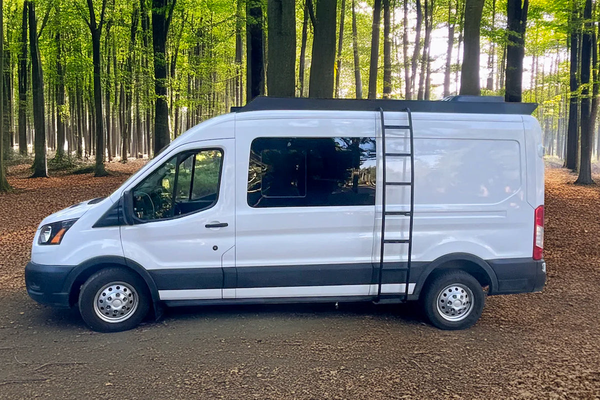 A white mid roof Ford Transit cargo van with orion van gear roof rack and side ladder parked on a dirt path in a forest with green trees and brown leaf litter on the ground.