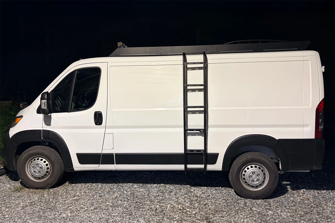 A white promaster cargo van parked outdoors at night on gravel, with a black orion side ladder attached vertically along the side door and a black orion stealth+roof rack visible above.