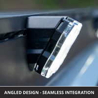 Close-up of a vehicle roof rack rock light mounted at an angle on a van roof rack, with a clear lens and visible internal reflector. Text at the bottom reads 'ANGLED DESIGN - SEAMLESS INTEGRATION'.