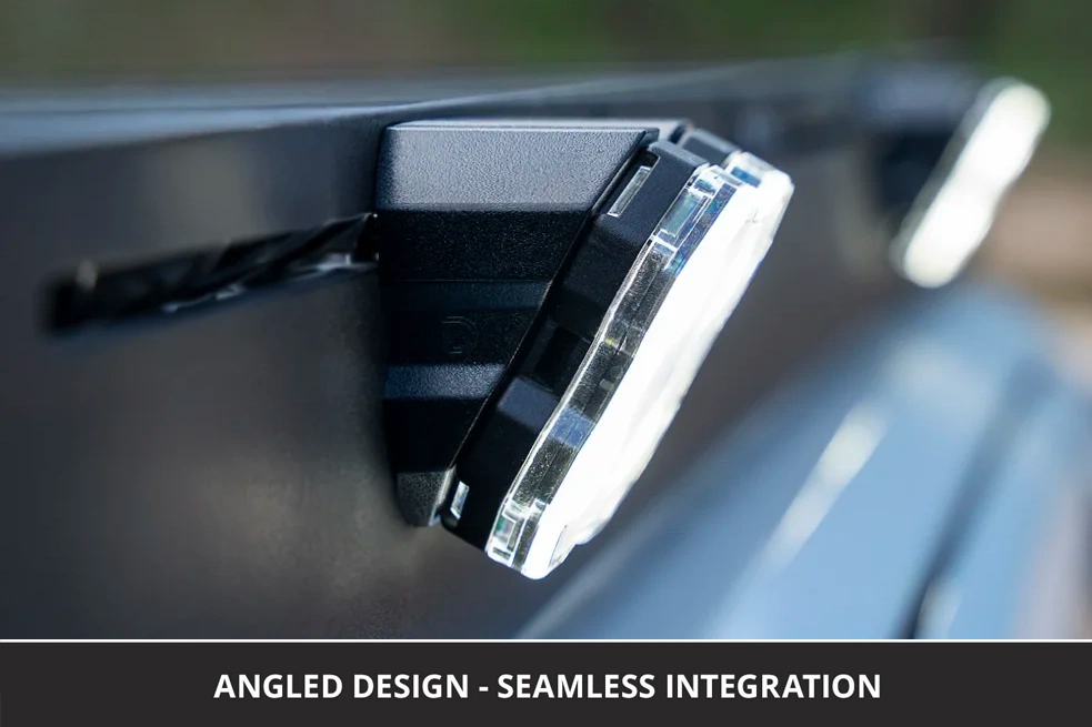 Close-up of a vehicle roof rack rock light mounted at an angle on a van roof rack, with a clear lens and visible internal reflector. Text at the bottom reads 'ANGLED DESIGN - SEAMLESS INTEGRATION'.