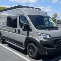 A gray Ram ProMaster camper van with orion roof rack parked in an outdoor parking lot under a blue sky with trees in the background.