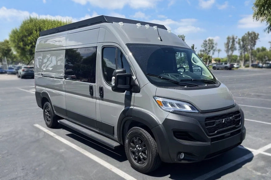 A gray Ram ProMaster camper van with orion roof rack parked in an outdoor parking lot under a blue sky with trees in the background.