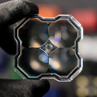 A hand wearing a black glove holds a square, multi-lens optical glass filter or lens assembly with a silver metallic frame against a dark, blurred background.