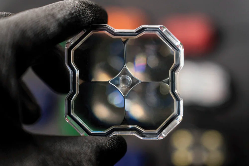 A hand wearing a black glove holds a square, multi-lens optical glass filter or lens assembly with a silver metallic frame against a dark, blurred background.