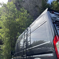 A ram promaster 159 van parked outdoors in a wooded area with a large rocky cliff behind it and orion van gear roof rack and side ladder