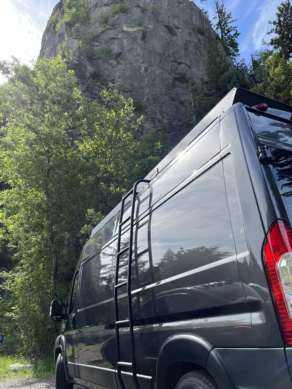 A ram promaster 159 van parked outdoors in a wooded area with a large rocky cliff behind it and orion van gear roof rack and side ladder