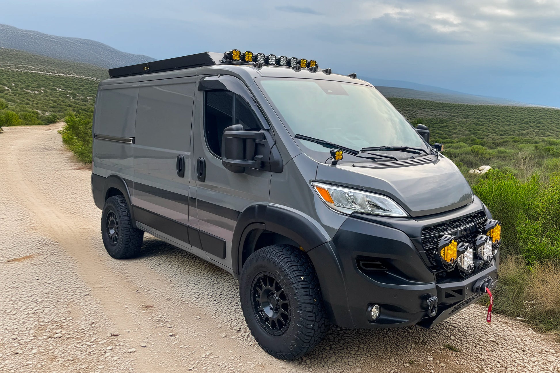 Gray promaster low roof 118 van with off-road modifications on a dirt road with a scenic background